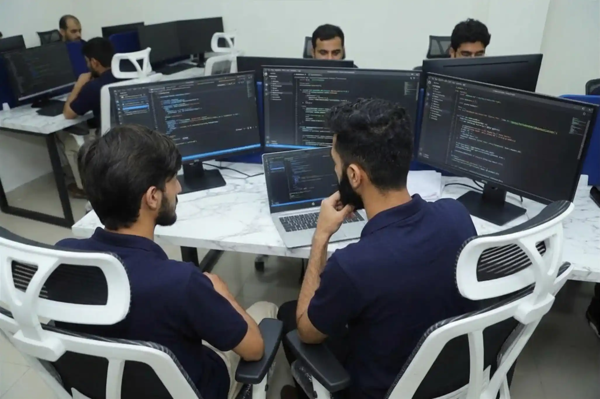 Two software developers collaborating at a marble desk with multiple monitors displaying code in a modern, minimalist tech office.